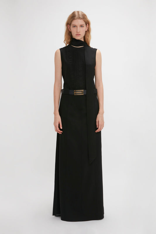 Person standing against a white background, wearing a Victoria Beckham Tailored Floor-Length Skirt In Black with a high neck and a belt, featuring a structured silhouette.