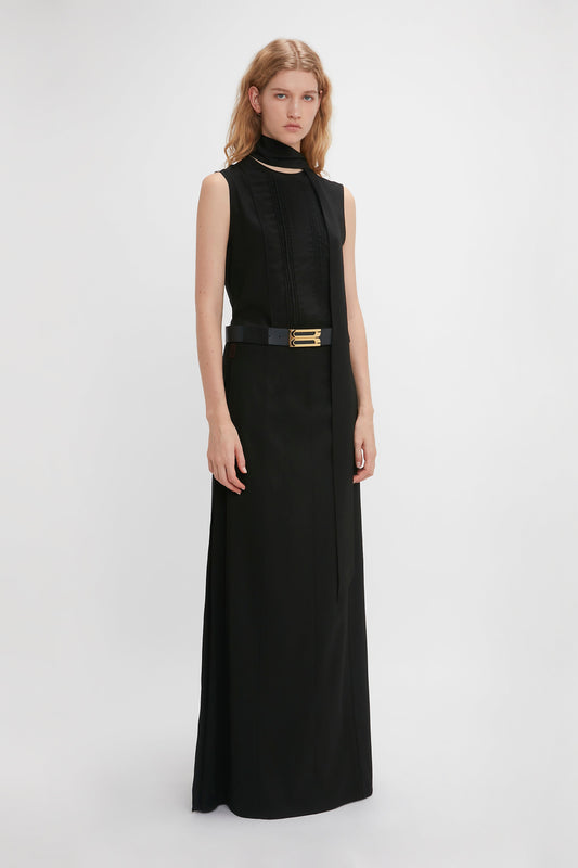 A person with long hair is wearing a sleeveless black dress with a high neckline, a belt, and a structured silhouette. They are standing against a plain white background, showcasing the Victoria Beckham Tailored Floor-Length Skirt In Black.