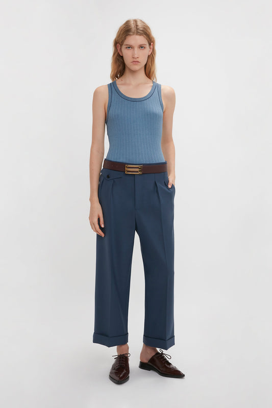 A person with long hair is wearing a Victoria Beckham Fine Knit Micro Stripe Tank In Heritage Blue, dark blue trousers with a brown belt, and brown shoes, standing against a plain white background.