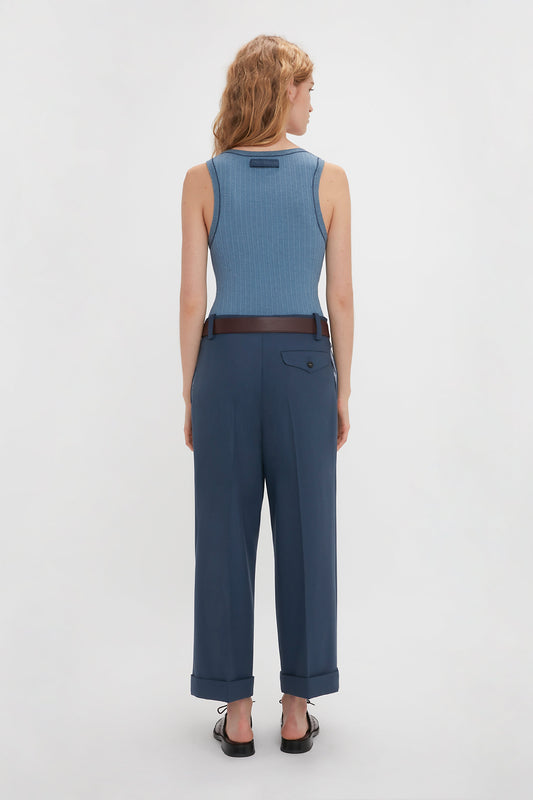 A person with wavy blonde hair is standing with their back facing the camera, wearing a Victoria Beckham Fine Knit Micro Stripe Tank In Heritage Blue, high-waisted navy blue pants, and black slip-on shoes reminiscent of Victoria Beckham's sleek style.