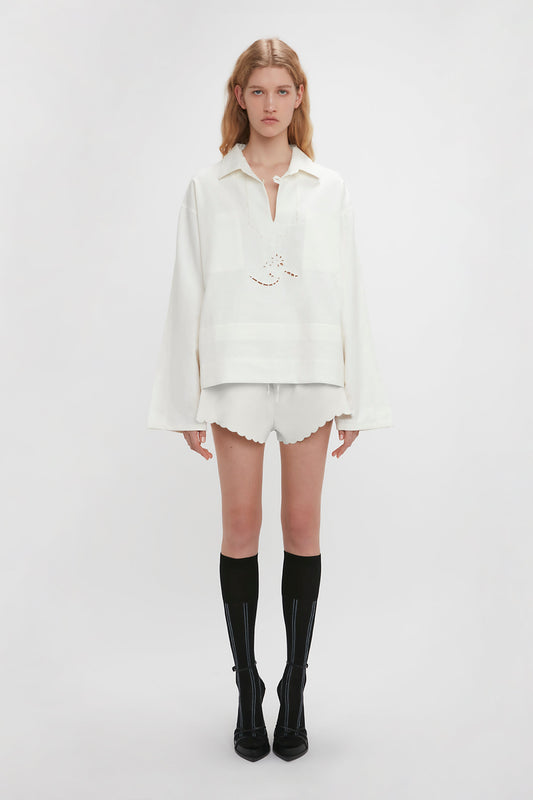 A person stands against a plain background in Victoria Beckham’s runway picks: an Oversized Embroidered Tunic In Antique White, white scalloped shorts, knee-high black socks, and black shoes. Their blonde hair falls neatly, complementing their neutral expression.