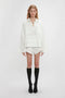 A person stands against a plain background in Victoria Beckham’s runway picks: an Oversized Embroidered Tunic In Antique White, white scalloped shorts, knee-high black socks, and black shoes. Their blonde hair falls neatly, complementing their neutral expression.