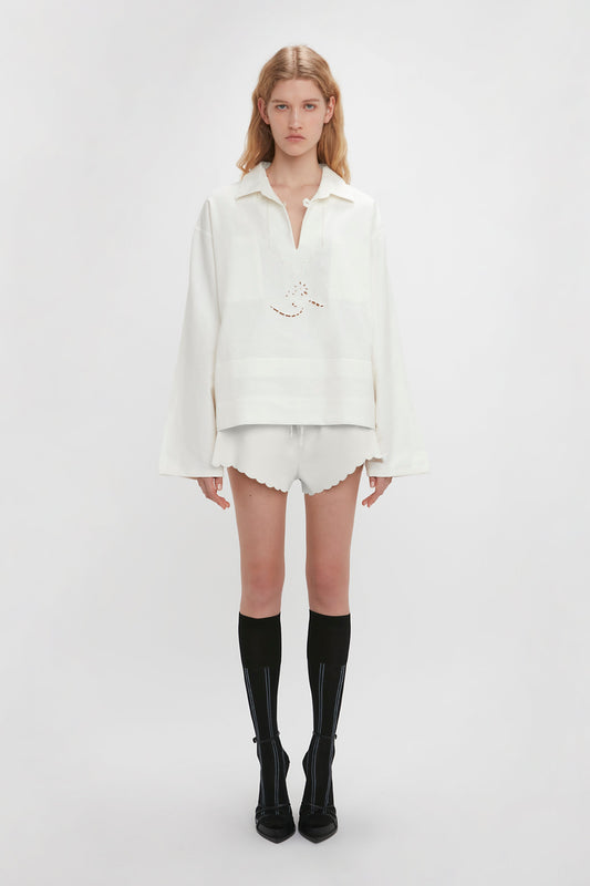 A person stands against a plain background wearing a white blouse with delicate embroidered detailing, Drawstring Embroidered Mini Short In Antique White by Victoria Beckham, and knee-high black socks with black shoes.