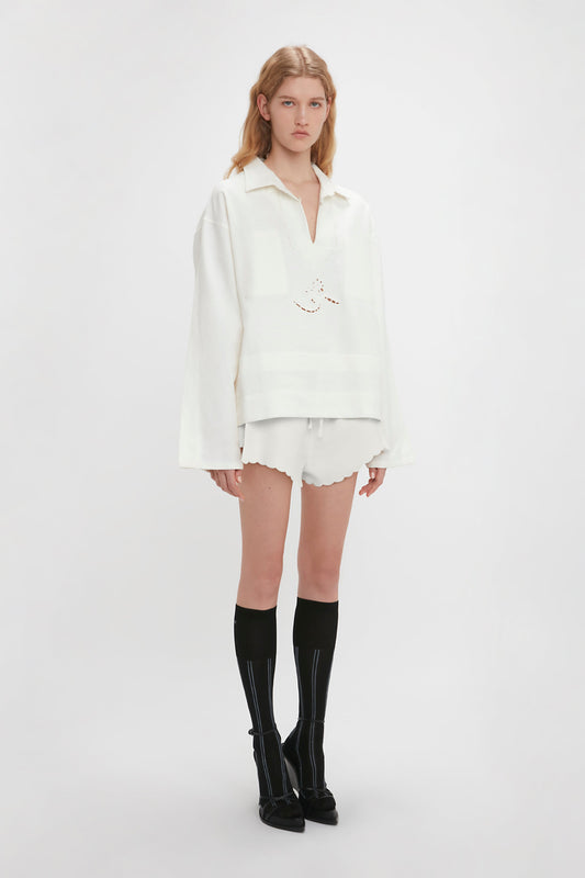 A person stands against a plain background, showcasing Victoria Beckham’s runway picks: an oversized Embroidered Tunic In Antique White paired with white shorts and black knee-high socks with black shoes. They have long, light brown hair.