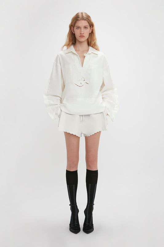 A woman wearing a white top with embroidered detailing and Victoria Beckham's Drawstring Embroidered Mini Short In Antique White stands against a plain white background. Black knee-high socks and black heels add contrast to her vintage appeal.