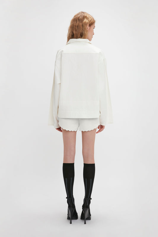 A person with long hair is standing facing away, wearing an Oversized Embroidered Tunic In Antique White by Victoria Beckham, complemented by black knee-high socks with high-heeled shoes on a white background. This ensemble could easily be featured among Victoria’s runway picks.