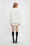 A person with long hair is standing facing away, wearing an Oversized Embroidered Tunic In Antique White by Victoria Beckham, complemented by black knee-high socks with high-heeled shoes on a white background. This ensemble could easily be featured among Victoria’s runway picks.