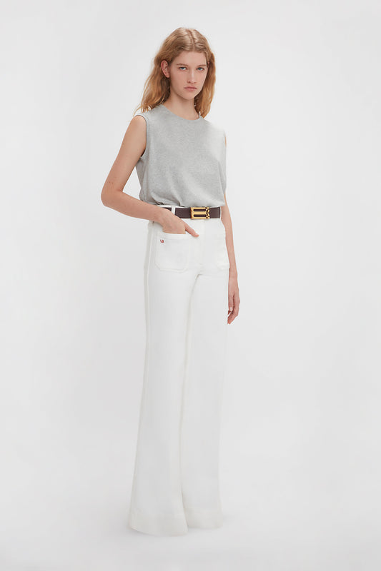 A person with long hair wearing a versatile *Sleeveless T-Shirt In Grey Marl* by *Victoria Beckham*, white high-waisted pants made of organic cotton, and a dark belt stands against a plain white background.