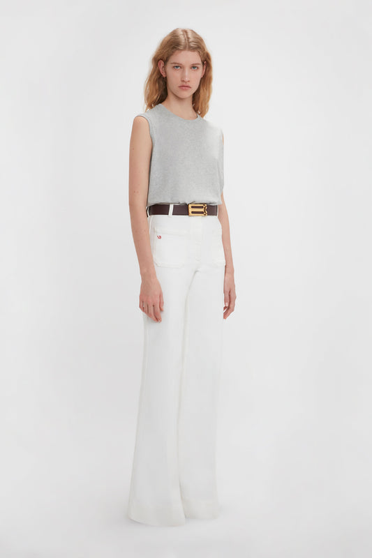 A woman stands against a white background wearing a versatile Victoria Beckham Sleeveless T-Shirt In Grey Marl, white wide-leg pants, and a black belt with a gold buckle.