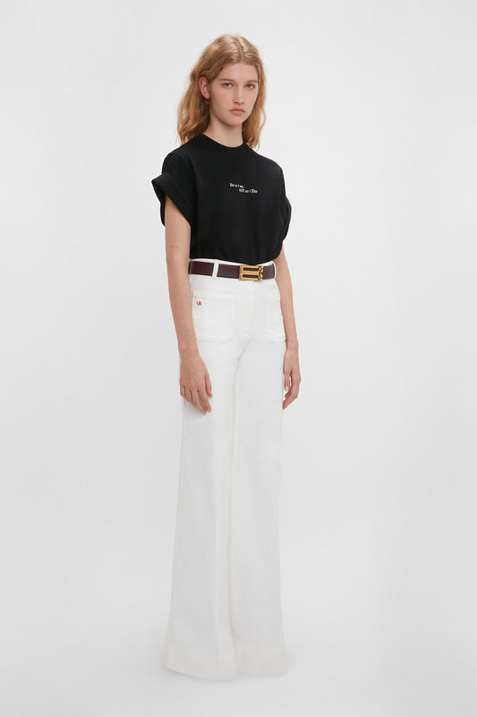 A woman in a Victoria Beckham 'Do As I Say, Not As I Do' Slogan T-shirt in black and white high-waisted trousers, accessorized with a gold belt, poses against a plain white background.
