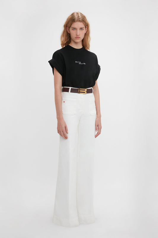 Young woman in a Victoria Beckham 'Do As I Say, Not As I Do' Slogan T-shirt and white high-waisted flared jeans, standing against a plain white background.