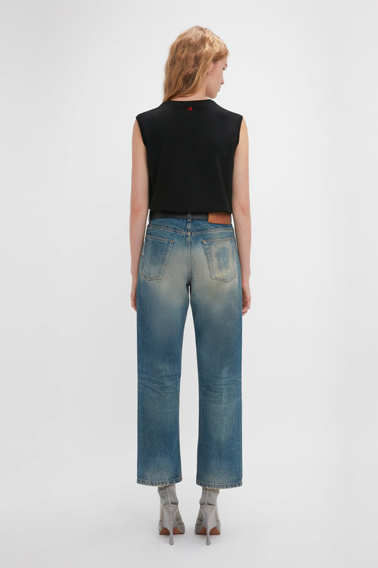 Woman in black sleeveless top and Victoria Beckham heritage twill denim jeans with a Relaxed Straight Leg Jean In Antique Indigo Wash, standing with her back to the camera on a white background.