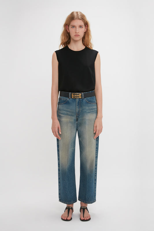A woman with long hair wears a sleeveless black top tucked into blue jeans with frayed hems and Victoria Beckham's Flat Chain Sandal In Black Leather. She also has a black belt with a gold buckle. The background is plain white, showcasing the effortless style of the SS24 collection.