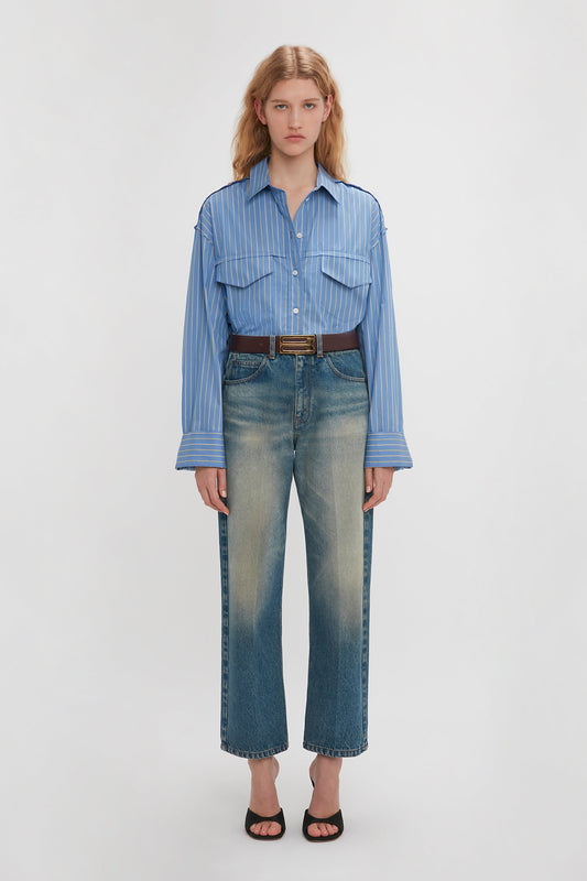 A person stands against a plain background, wearing a Victoria Beckham Cropped Seam Detail Shirt In Steel Blue, faded jeans, and black open-toed shoes.