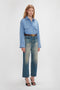 A person with long hair stands wearing a Cropped Seam Detail Shirt In Steel Blue by Victoria Beckham, belted faded jeans, and black heeled sandals against a plain white background.