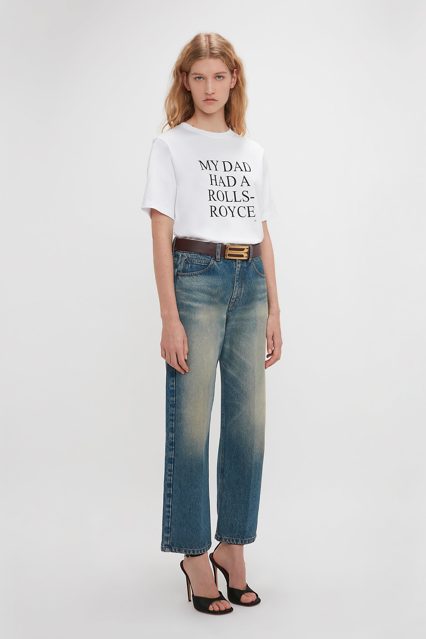 A woman standing in a studio wearing a white Exclusive 'My Dad Had A Rolls-Royce' slogan T-shirt by Victoria Beckham and faded blue jeans, paired with black heels.