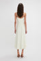 A woman with long hair is standing facing away, wearing an Exclusive Panelled Midi Cami Dress In Vanilla by Victoria Beckham and beige high heels.