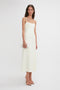 A woman in an Exclusive Panelled Midi Cami Dress In Vanilla by Victoria Beckham stands against a plain white background, holding a small, white clutch. She is also wearing nude-colored, open-toe heels.