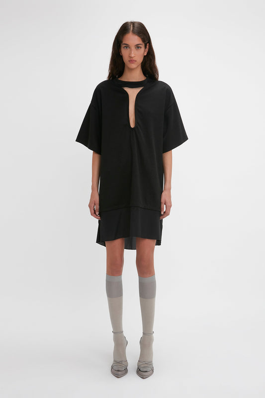 A woman stands against a plain white background, wearing a Frame Cut-Out T-Shirt Dress In Black by Victoria Beckham, gray knee-high socks, and gray pointed shoes. She has long dark hair and a neutral expression.