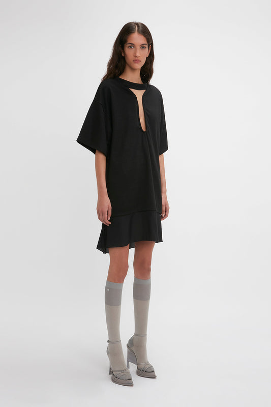 A woman stands against a plain white background, wearing the Victoria Beckham Frame Cut-Out T-Shirt Dress In Black, paired with knee-high grey socks and silver pointed-toe heels.