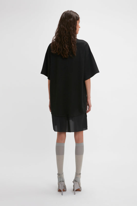 A person with long hair stands facing away from the camera, wearing a loose-fitting black Frame Cut-Out T-Shirt Dress In Black by Victoria Beckham, a black skirt, grey knee-high socks, and grey high-heeled shoes. The background is plain white.