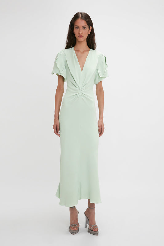 A woman in a mint green Victoria Beckham Gathered V-Neck Midi Dress In Jade with short sleeves and a waist-defining pleat, standing against a plain white background.