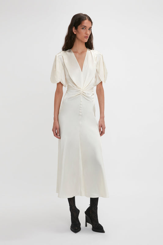 Woman wearing a Victoria Beckham Gathered V-Neck Midi Dress In Ivory with puffed short sleeves and waist-defining pleats, paired with black high-heeled shoes, standing against a plain white background.