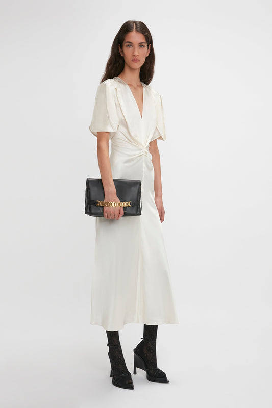 A person stands against a plain background wearing a Victoria Beckham Gathered V-Neck Midi Dress In Ivory with black ankle socks featuring lace detail, black high heels, and holding a black clutch bag with gold lettering.