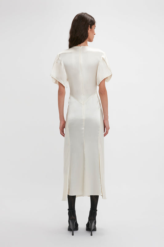 Rear view of a person with long dark hair wearing a Victoria Beckham Gathered V-Neck Midi Dress In Ivory with short sleeves and black high-heeled boots, standing against a plain white background.