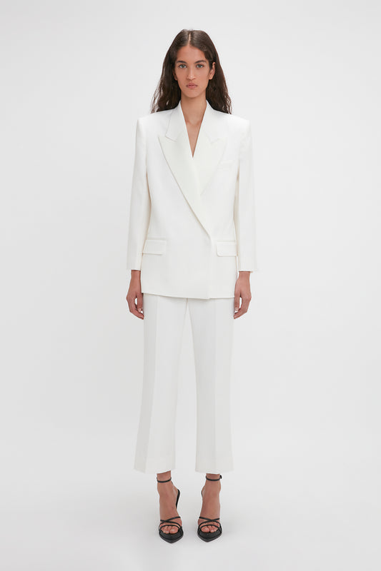 A person stands against a plain white background, wearing a white tailored pantsuit with Victoria Beckham Exclusive Cropped Tuxedo Trouser In Ivory and black strappy heels.