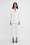 A person stands against a plain white background, wearing a white tailored pantsuit with Victoria Beckham Exclusive Cropped Tuxedo Trouser In Ivory and black strappy heels.