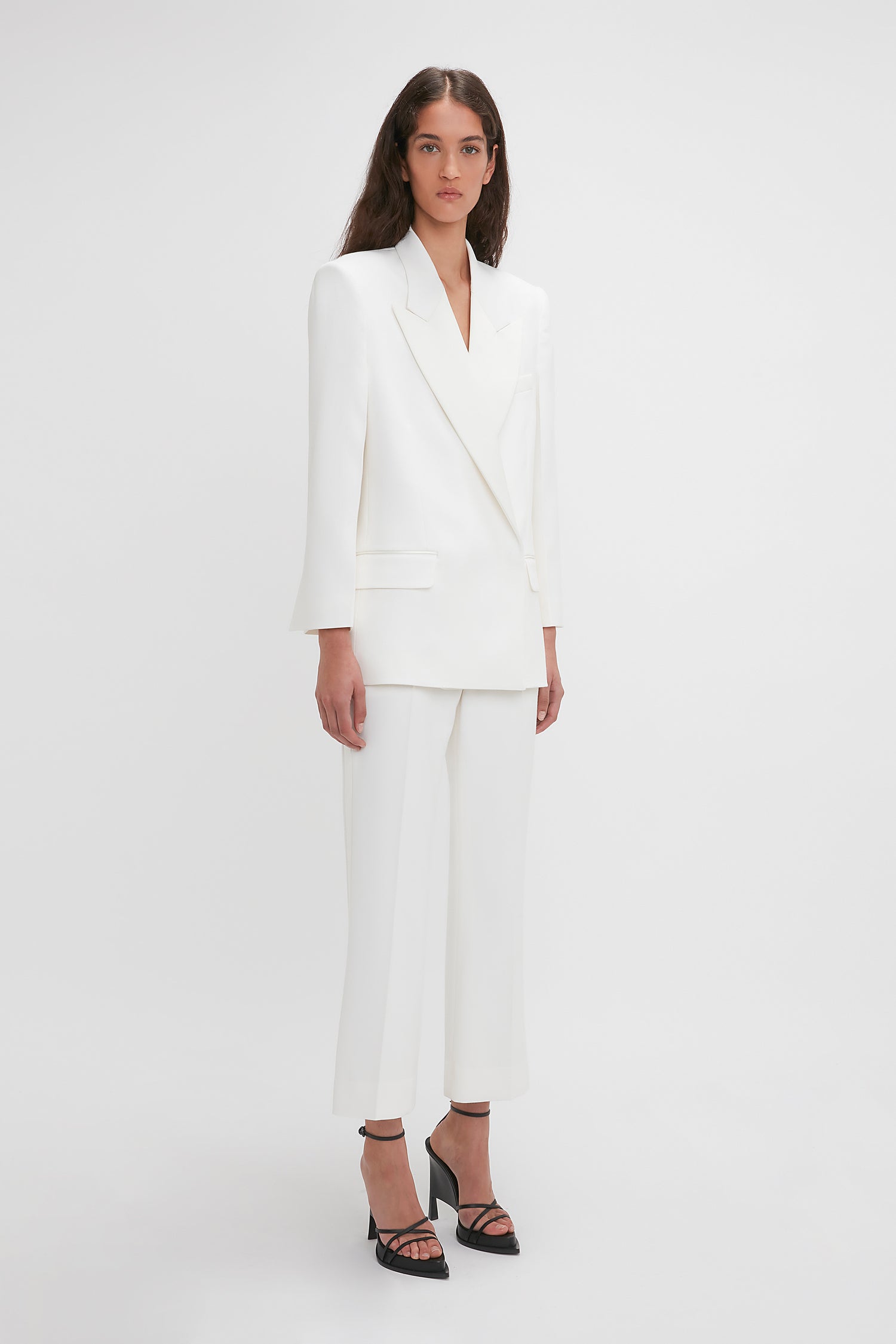 A person stands against a plain background wearing a white tailored suit featuring Victoria Beckham's Exclusive Cropped Tuxedo Trouser In Ivory with wide-legged pants and black platform heels.