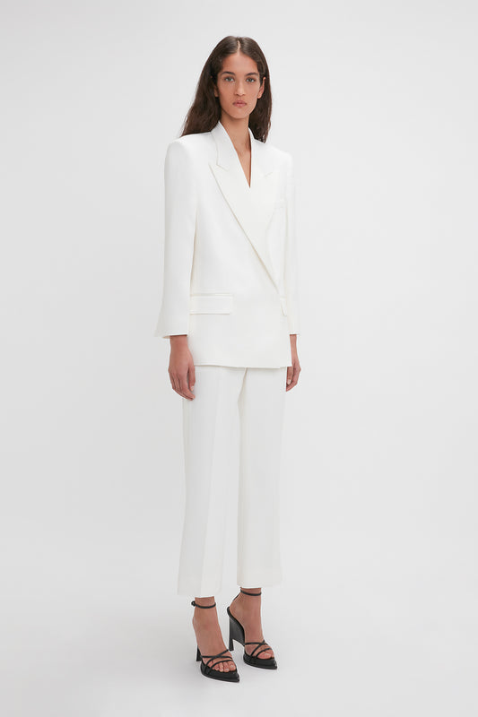 A person stands against a plain background wearing a white tailored suit featuring Victoria Beckham's Exclusive Cropped Tuxedo Trouser In Ivory with wide-legged pants and black platform heels.