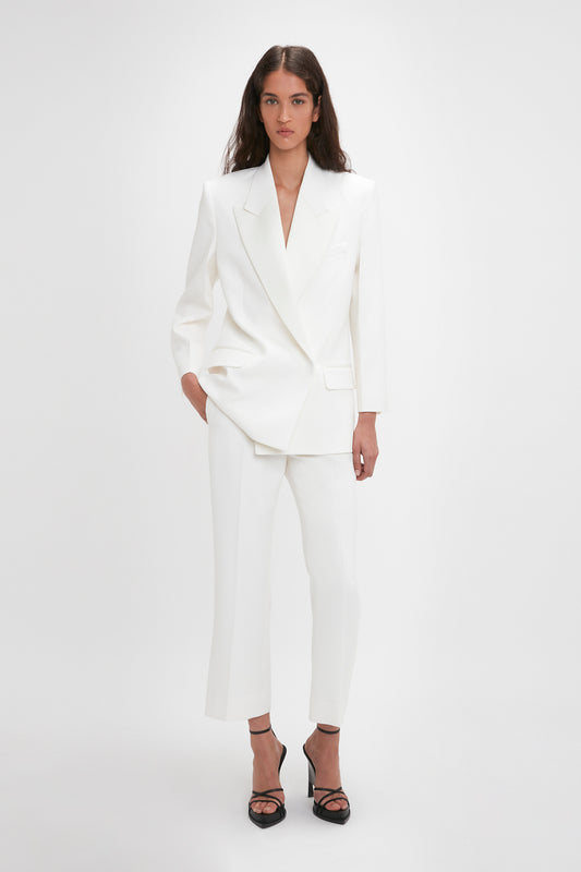 A woman stands against a plain background wearing a white blazer with matching Victoria Beckham Exclusive Cropped Tuxedo Trouser In Ivory and black high-heeled shoes. One hand is in her pocket, and she looks directly at the camera.