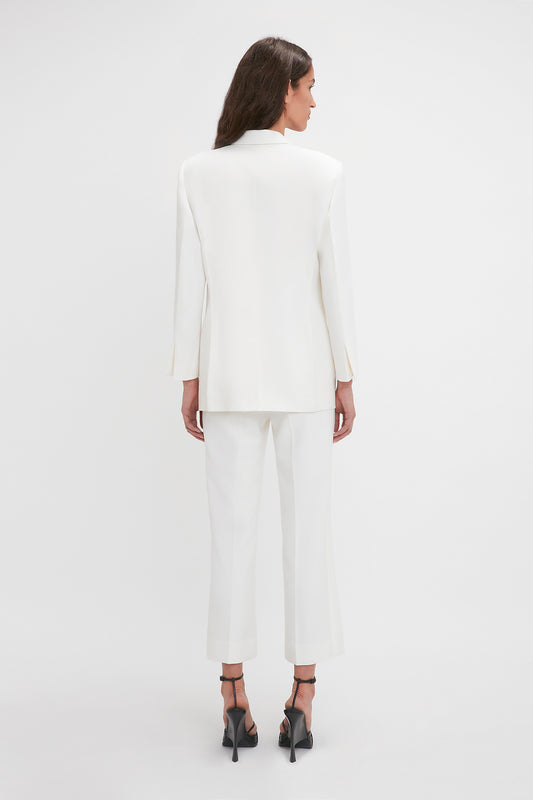 A person with long brown hair wearing a white blazer and Exclusive Cropped Tuxedo Trouser In Ivory by Victoria Beckham is standing facing away from the camera.
