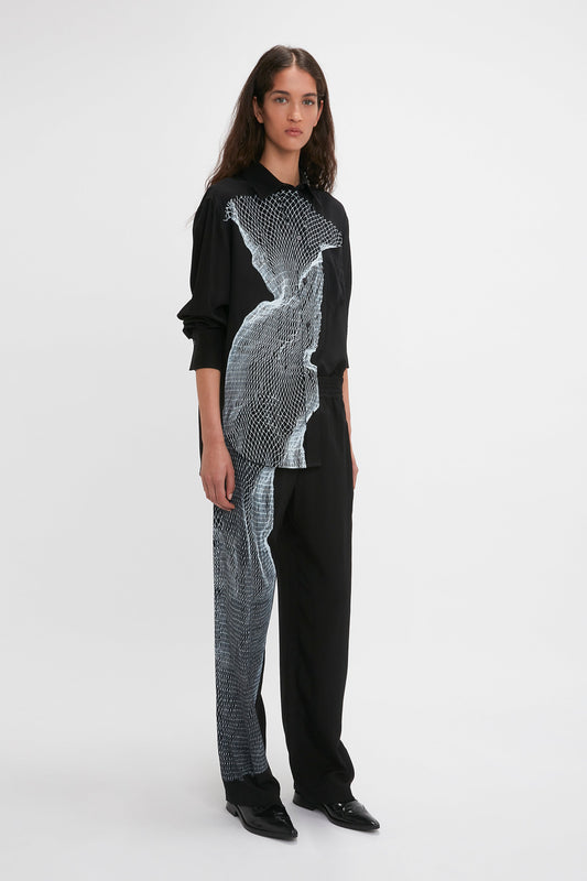 A person with long brown hair is standing against a plain white background, wearing a black outfit featuring a white abstract pattern and a relaxed-fit Victoria Beckham Long Sleeve Pyjama Shirt In Black-White Contorted Net.