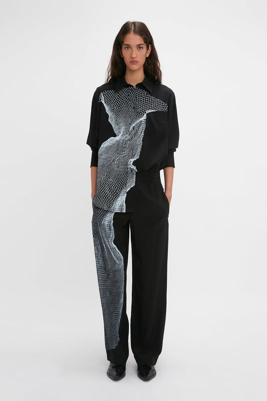 A person standing against a white background, wearing the Long Sleeve Pyjama Shirt In Black-White Contorted Net by Victoria Beckham, featuring an abstract graphic twisted net print. The person has long dark hair and is looking directly at the camera.