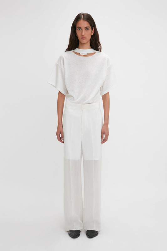 A person in a white short-sleeve top and Victoria Beckham Waistband Detail Straight Leg Trouser In White stands against a plain white background, looking forward.