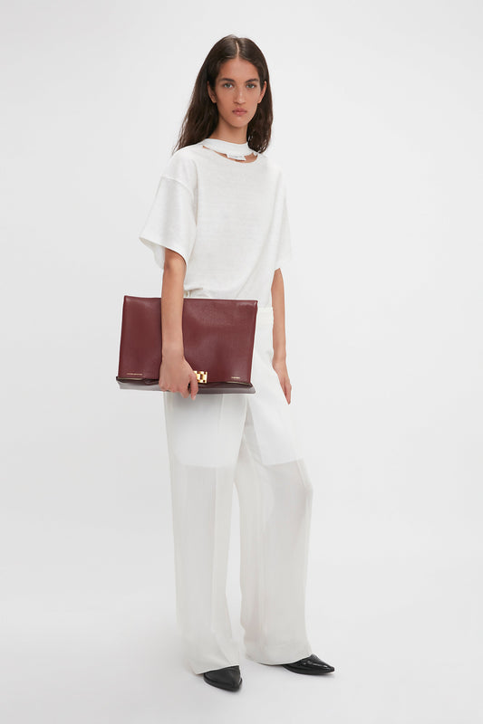 A woman in a loose white top and Victoria Beckham Waistband Detail Straight Leg Trouser In White holds a large maroon clutch. She is wearing black pointed shoes, standing against a plain white background, embodying contemporary cool.