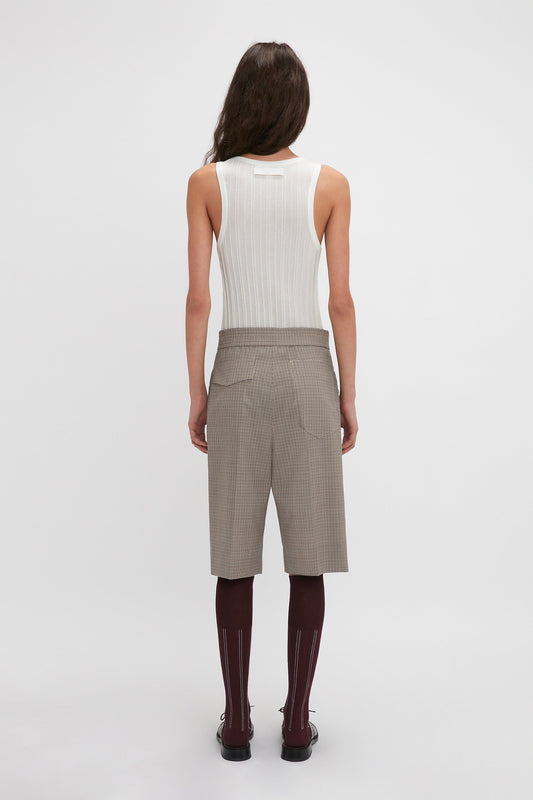A person with long hair is facing away from the camera, wearing a white sleeveless top, Victoria Beckham Waistband Detailed Tailored Short In Multi, and burgundy knee-high socks.