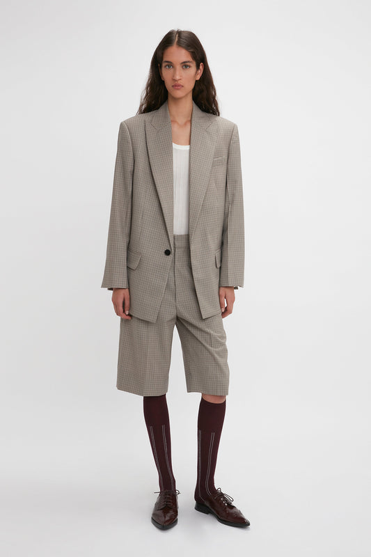 A person stands in a light-colored, oversized silhouette featuring the Victoria Beckham Peak Lapel Jacket In Multi and matching knee-length shorts, paired with maroon knee-high socks and brown shoes, with a neutral expression against a white background.