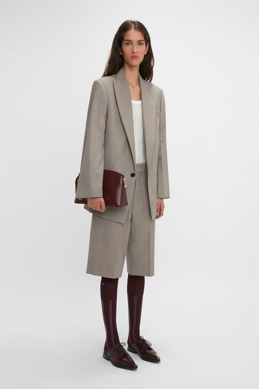 A woman stands against a plain background wearing a light-gray dogtooth check blazer made of virgin wool, Victoria Beckham Waistband Detailed Tailored Short In Multi, a white top, maroon socks, and dark brown shoes, holding a dark brown clutch.