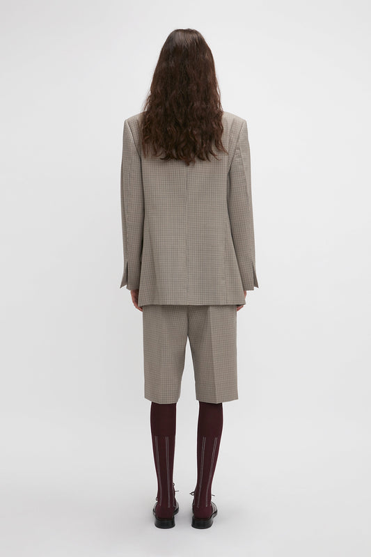 Back view of a person with long curly hair wearing a beige checkered Peak Lapel Jacket In Multi by Victoria Beckham featuring an oversized silhouette with knee-length shorts, long maroon socks, and black shoes, standing against a plain white background.