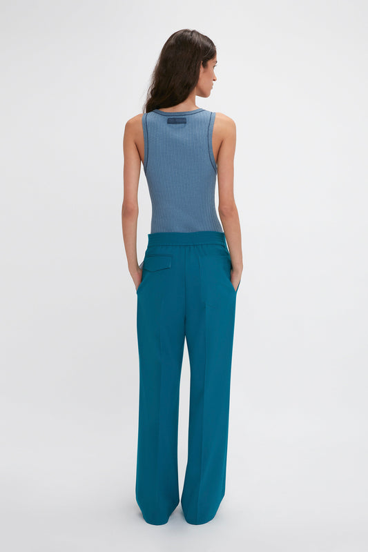 A woman with long brown hair is shown from the back wearing a sleeveless blue top and Waistband Detail Straight Leg Trouser In Petroleum by Victoria Beckham.