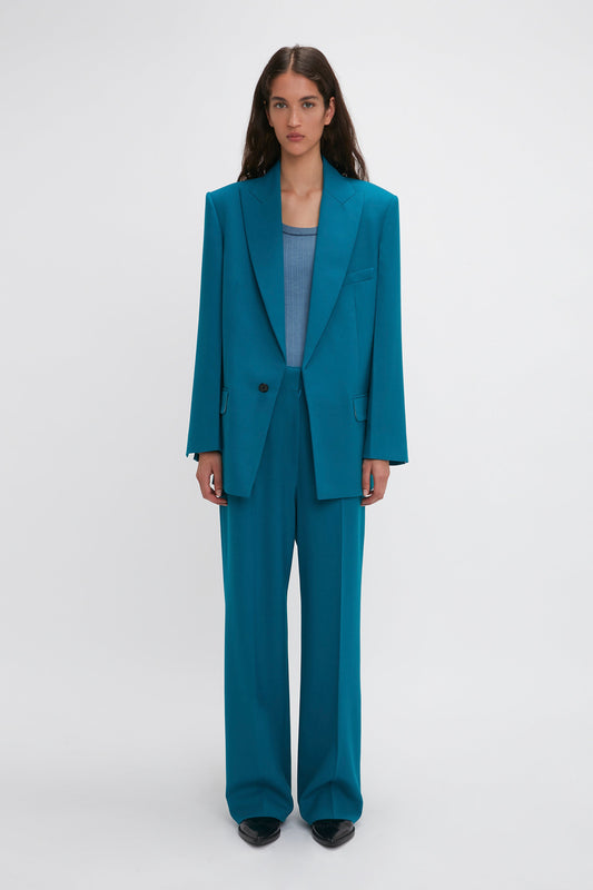 A person stands against a plain white background, wearing an oversized Peak Lapel Jacket In Petroleum from the Victoria Beckham brand with a matching teal top and black shoes.