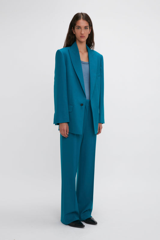A woman stands in front of a plain background wearing a Peak Lapel Jacket In Petroleum from the Victoria Beckham brand and a light blue top, paired with black shoes.