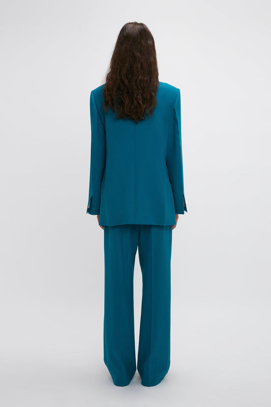 A person with long hair is standing facing away, wearing a teal suit by Victoria Beckham, featuring a Peak Lapel Jacket In Petroleum and wide-legged trousers that emphasize the oversized silhouette against a plain white background.