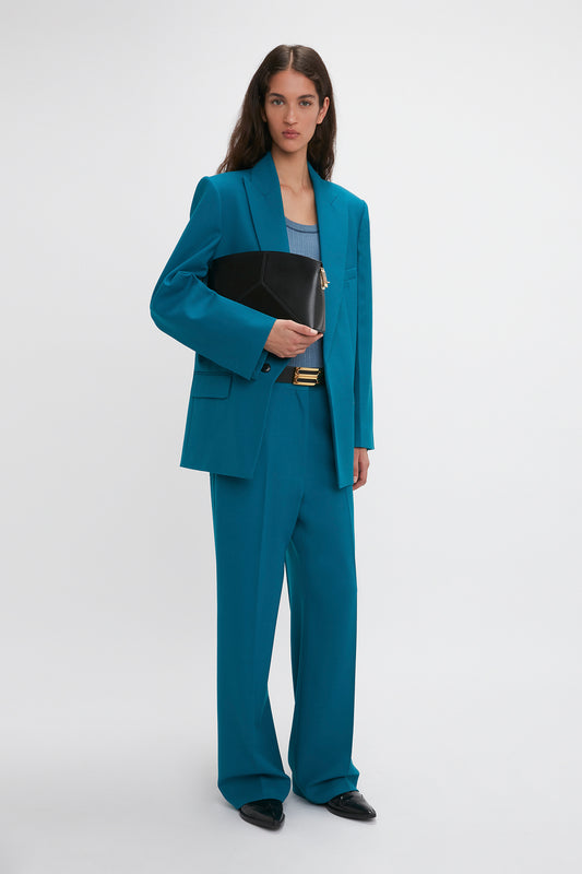 A person with long hair is standing against a plain background, wearing a teal suit featuring Victoria Beckham Waistband Detail Straight Leg Trouser In Petroleum and holding a black handbag.