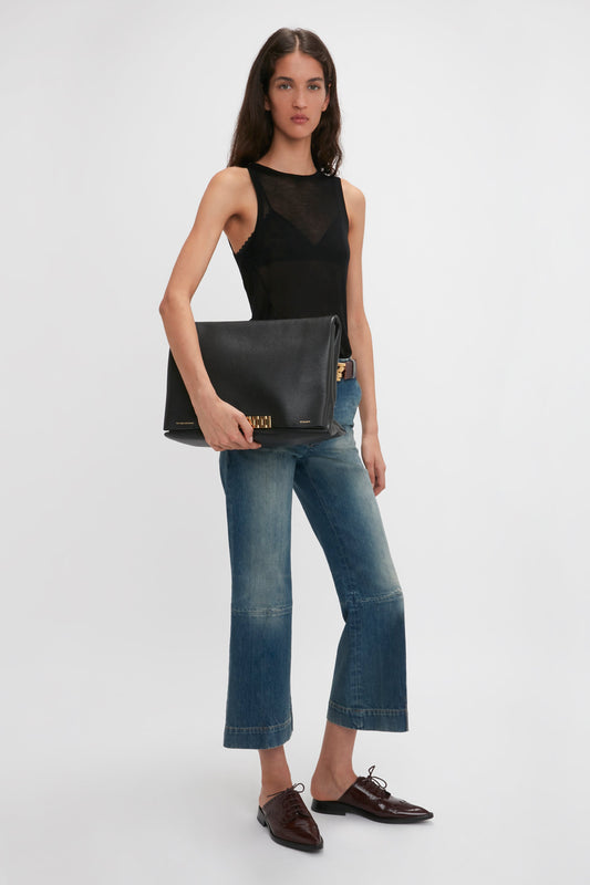 A person with long hair wearing a Lightweight Tank Top In Black by Victoria Beckham, blue jeans, and brown shoes holds a large black Givenchy clutch against a plain white background.