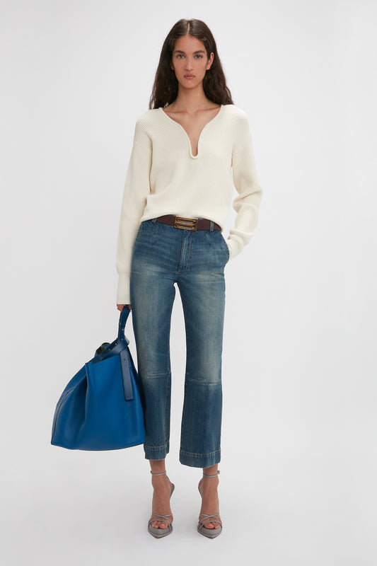 A woman wearing a white sweater, Victoria Beckham denim cropped kick jeans in Indigrey Wash, and holding a blue bag, standing against a plain background.